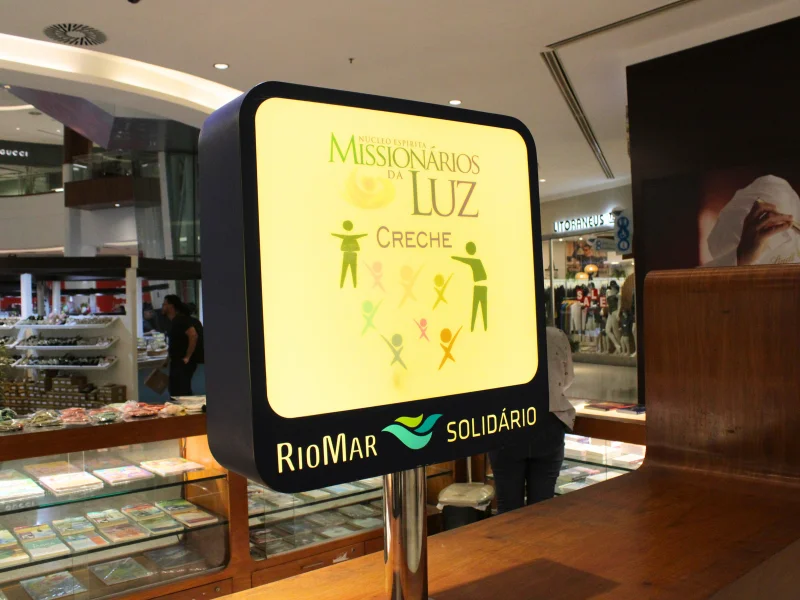 Close no totem luminoso do projeto "RioMar Solidário", destacando o logotipo do Núcleo Espírita Missionários da Luz e sua creche.