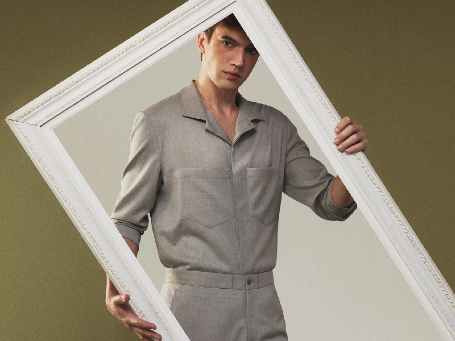 Jovem veste camisa cinza-claro com gola ampla e segura uma moldura de quadro branca, enquadrando-se nela. O fundo é de tonalidade neutra e suave. Jovem veste camisa cinza-claro com gola ampla e segura uma moldura de quadro branca, enquadrando-se nela. O fundo é de tonalidade neutra e suave.