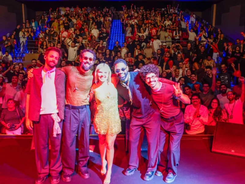 A cantora Duda Beat, de vestido curto verde, sorri no centro do palco abraçada a quatro músicos. Ao fundo, uma plateia lotada em um teatro levanta as mãos e segura pequenos balões vermelhos.