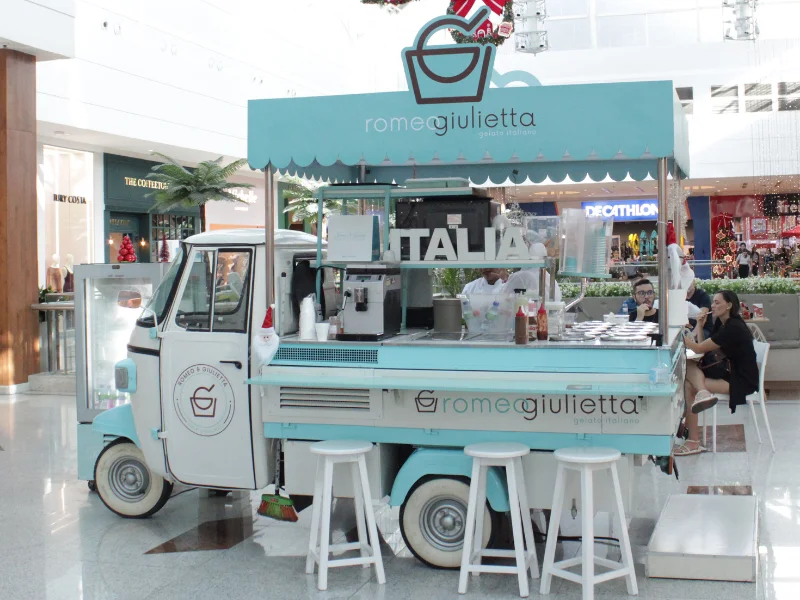 Um food truck azul e branco em formato de motoneta (Piaggio Ape), da sorveteria "Romeo e Giulietta Gelato Italiano", está em um corredor de shopping. Funcionários preparam os gelatos enquanto clientes sentam em banquetas brancas à direita.