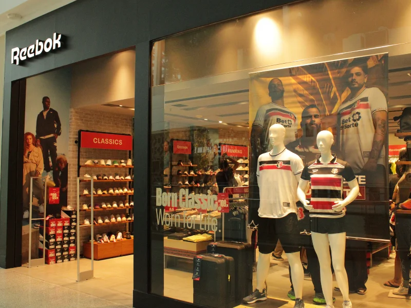Visão ampla da fachada da loja Reebok em um shopping, destacando o banner da campanha com jogadores do time.