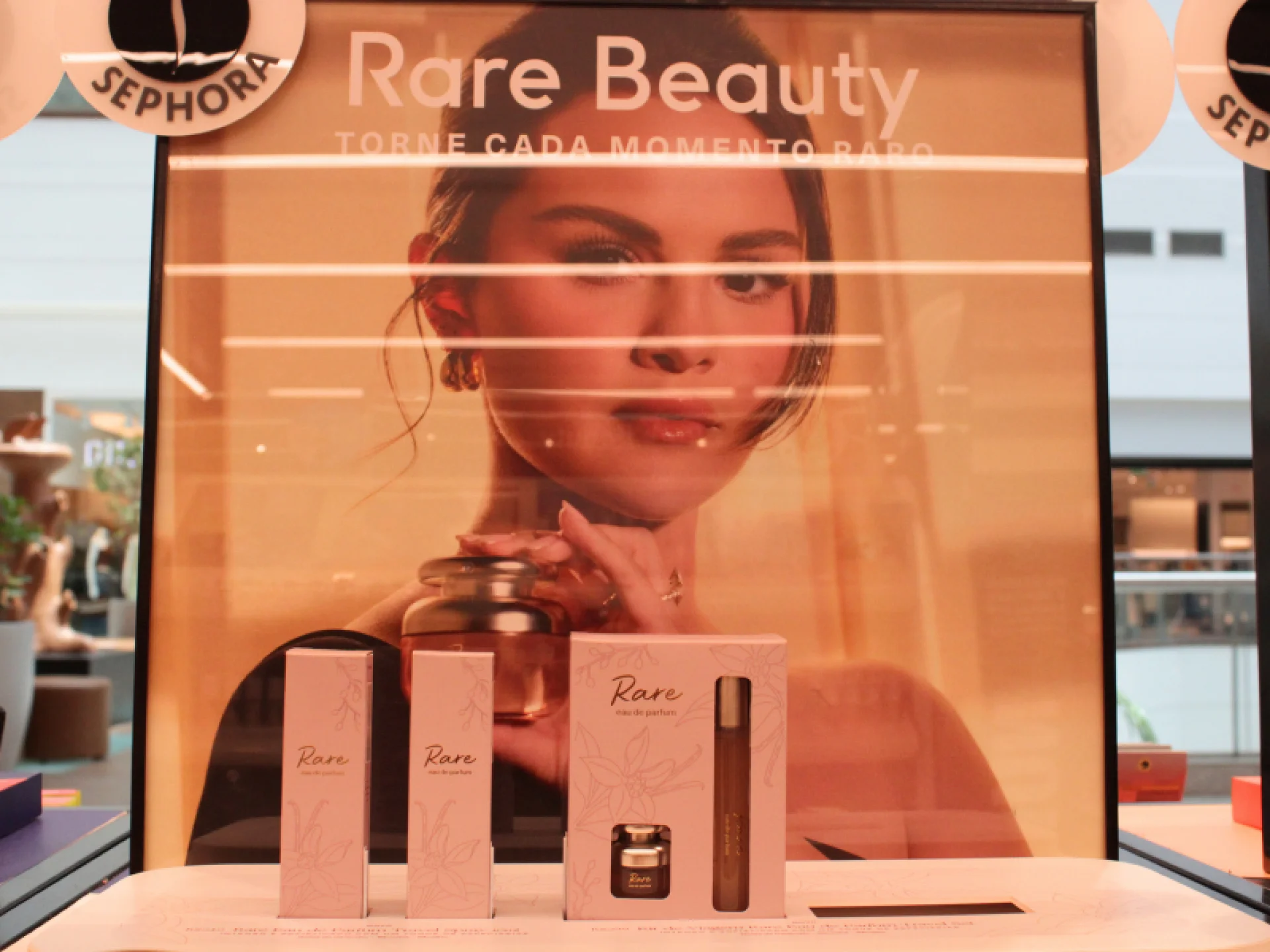 A imagem mostra a exibição do perfume "Rare Eau de Parfum" da Rare Beauty, com um display grande de Selena Gomez e as caixas dos produtos em miniatura. A imagem mostra a exibição do perfume "Rare Eau de Parfum" da Rare Beauty, com um display grande de Selena Gomez e as caixas dos produtos em miniatura.