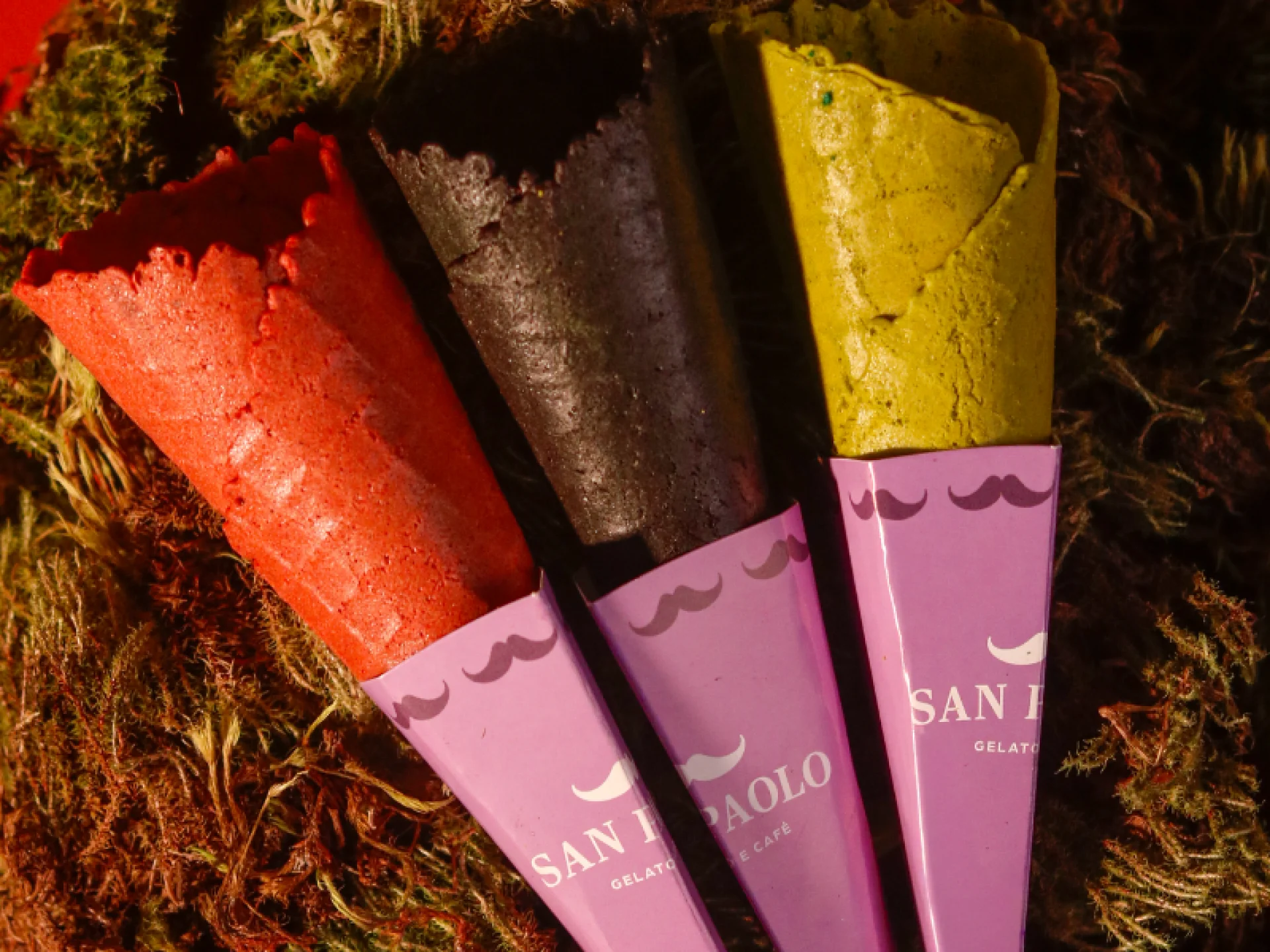 A imagem mostra três casquinhas de sorvete da San Paolo com sabores ou cores especiais: Coral, Preta e Verde, com embalagens personalizadas de bigode. A imagem mostra três casquinhas de sorvete da San Paolo com sabores ou cores especiais: Coral, Preta e Verde, com embalagens personalizadas de bigode.