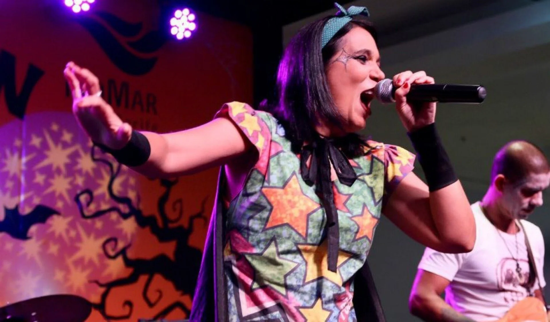 Uma mulher, vocalista da banda Mini Rock, canta intensamente em um palco com decoração de Halloween, segurando um microfone e vestindo uma roupa colorida de estrela.