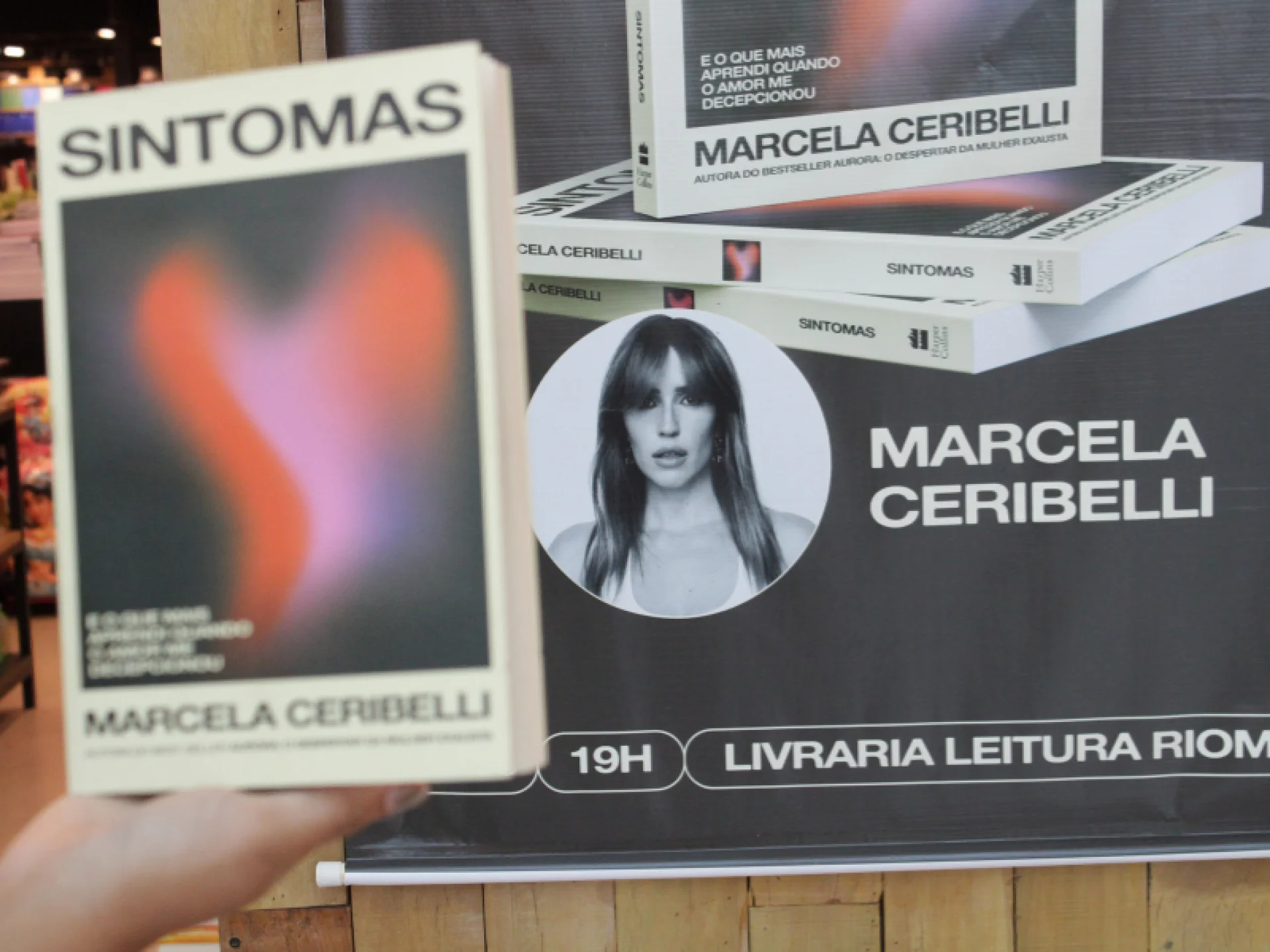 Um exemplar do livro "Sintomas" é segurado em frente a um banner do lançamento na Livraria Leitura do RioMar Recife. Um exemplar do livro "Sintomas" é segurado em frente a um banner do lançamento na Livraria Leitura do RioMar Recife.