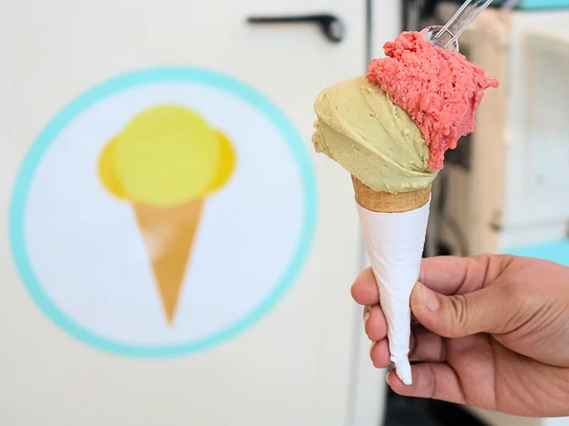 Mão segurando uma casquinha de sorvete com duas bolas, uma de cor creme e outra rosa avermelhada, à frente de um logotipo de sorvete na parede.