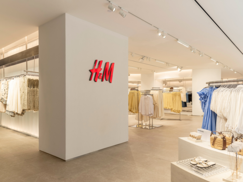Foto do interior da loja H&amp;M Iguatemi, com interior claro e uma parede com a logo da marca. Vemos, ainda, produtos expostos em araras e mesas.