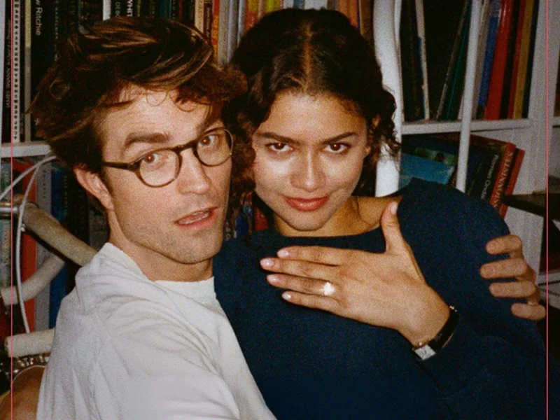 Um pôster granulado em estilo retrô mostra Robert Pattinson e Zendaya abraçados em uma biblioteca; ela exibe um anel brilhante enquanto ele olha surpreso para a câmera.
