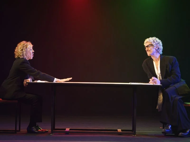 Dois atores estão sentados em extremidades opostas de uma mesa longa em um palco escuro. À esquerda, a mulher estende a mão sobre a mesa enquanto fala; à direita, o homem sorri com as mãos entrelaçadas.