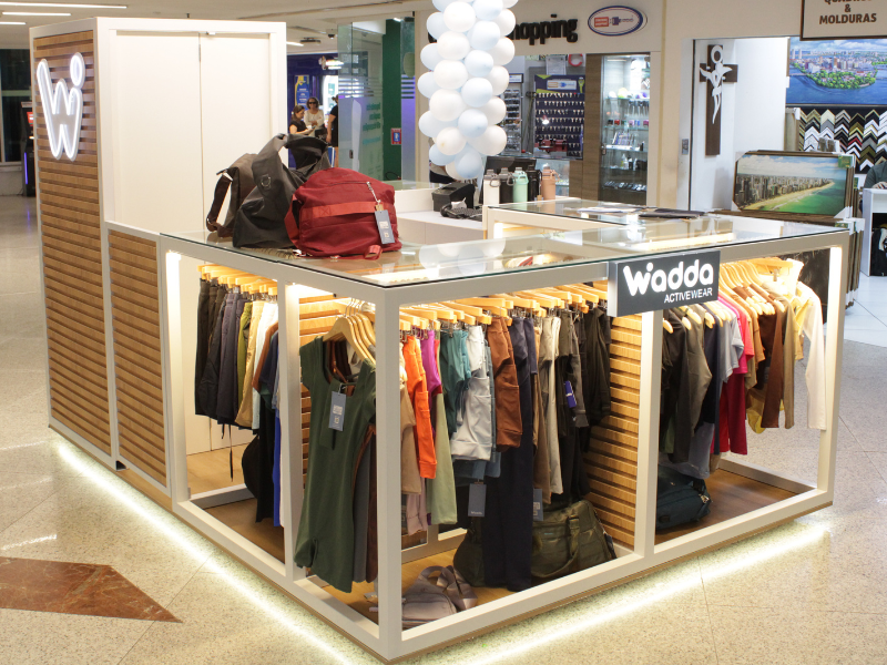 Visão geral do quiosque moderno da loja em um shopping, feito com estrutura branca, detalhes em madeira e araras cheias de roupas de treino.